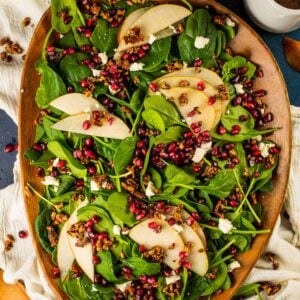 spinach salad on a brown wooden tray topped with pears, pomegranate, gorgonzola and pecans