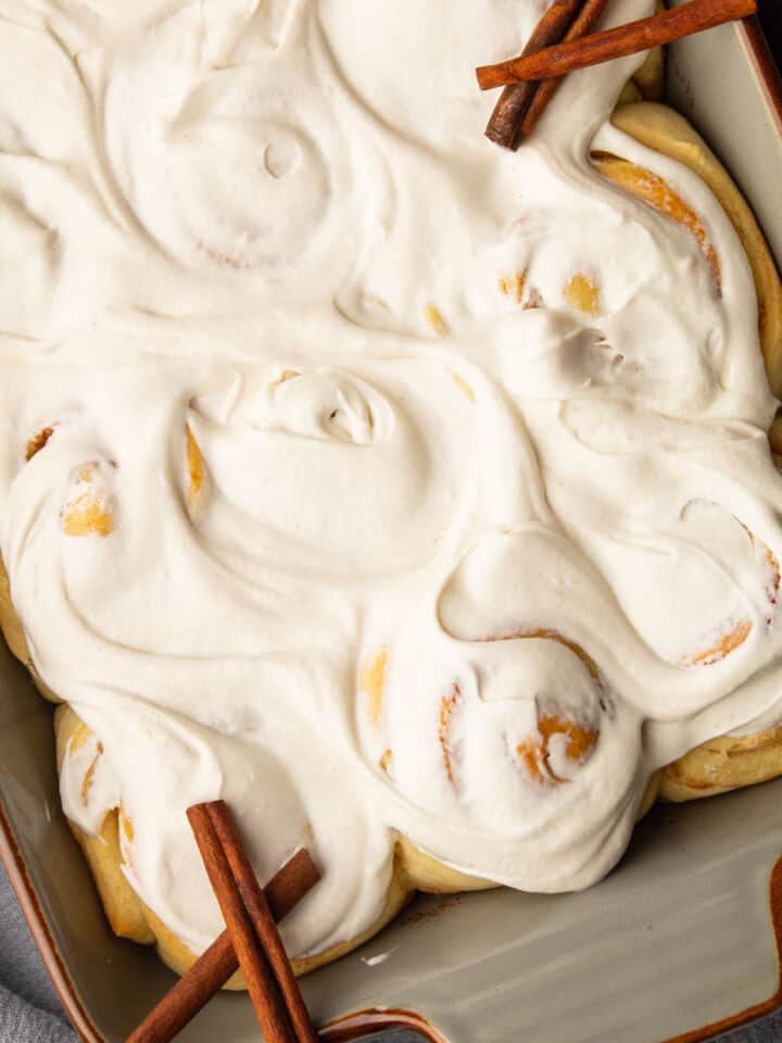 pan of cinnamon rolls covered in icing with a few cinnamon sticks around.