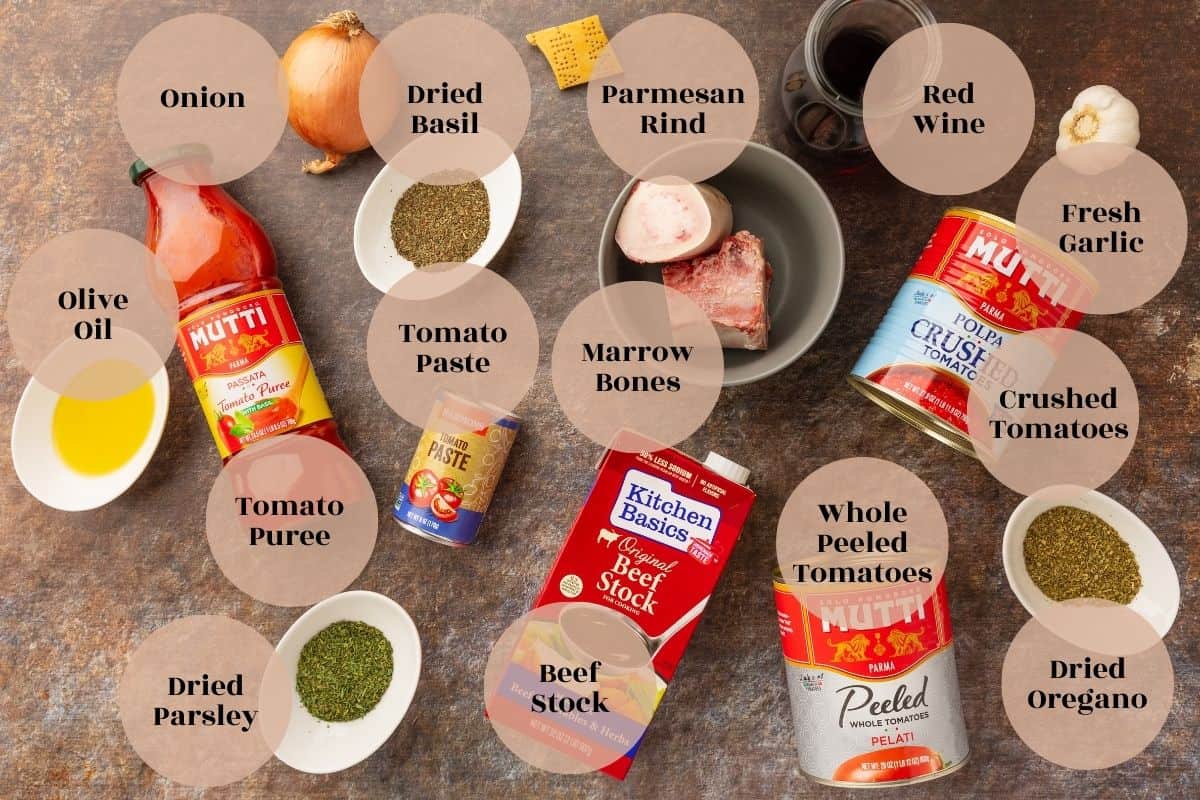 Cans of whole peeled and crushed tomatoes, carton of beef stock, can of tomato paste, jar of tomato purée, onion, garlic head, Parmesan rind, olive oil and dried herbs in dishes.