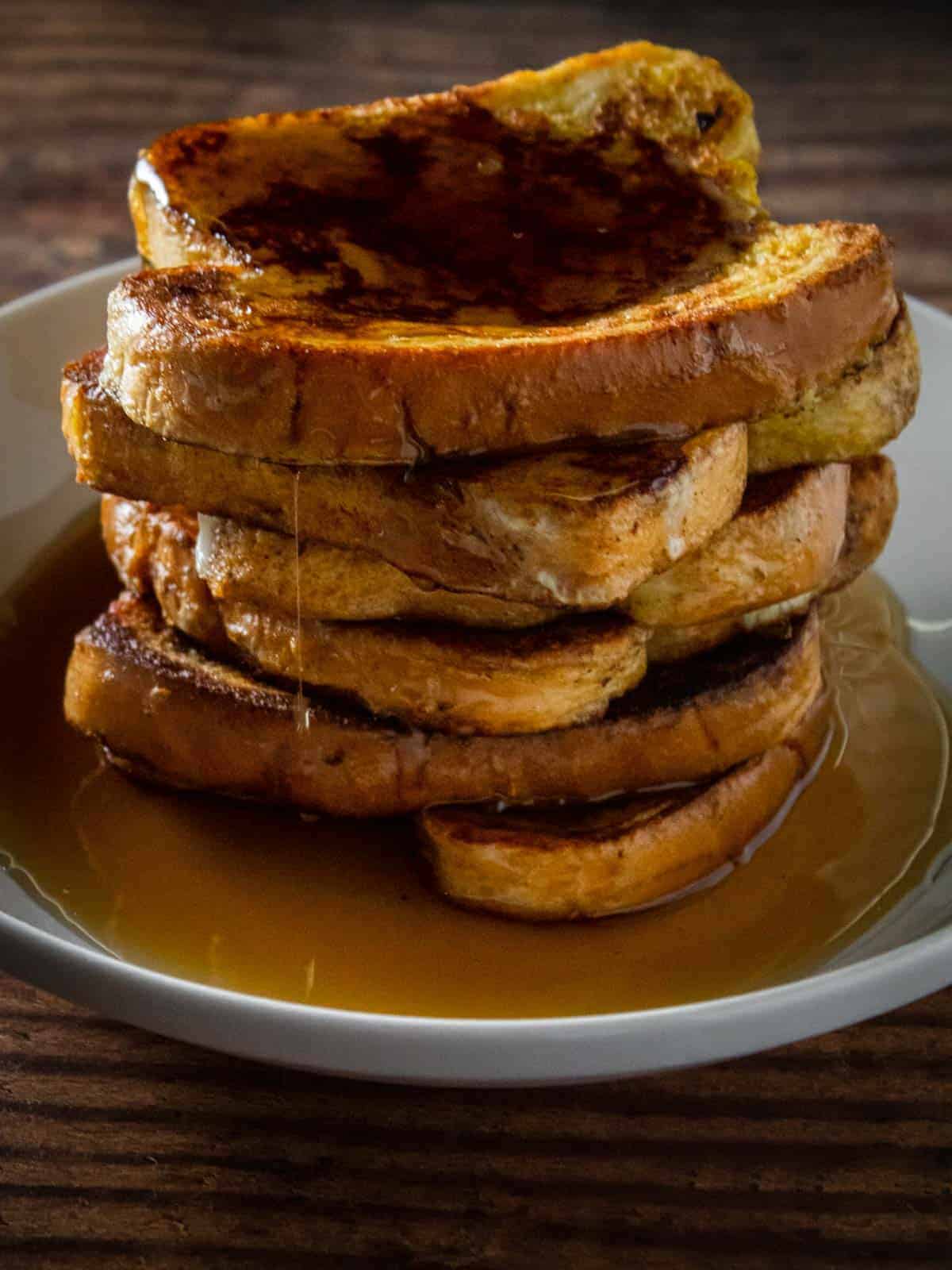 stack of french toast slices on a plate with syrup poured on it and dripping down the side.