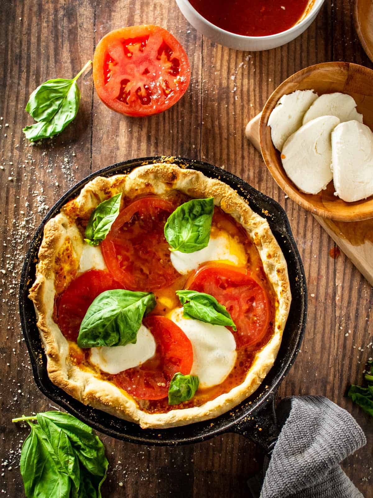 pizza in a cast iron skillet with tomatoes, cheese and fresh basil on it and in bowls around.