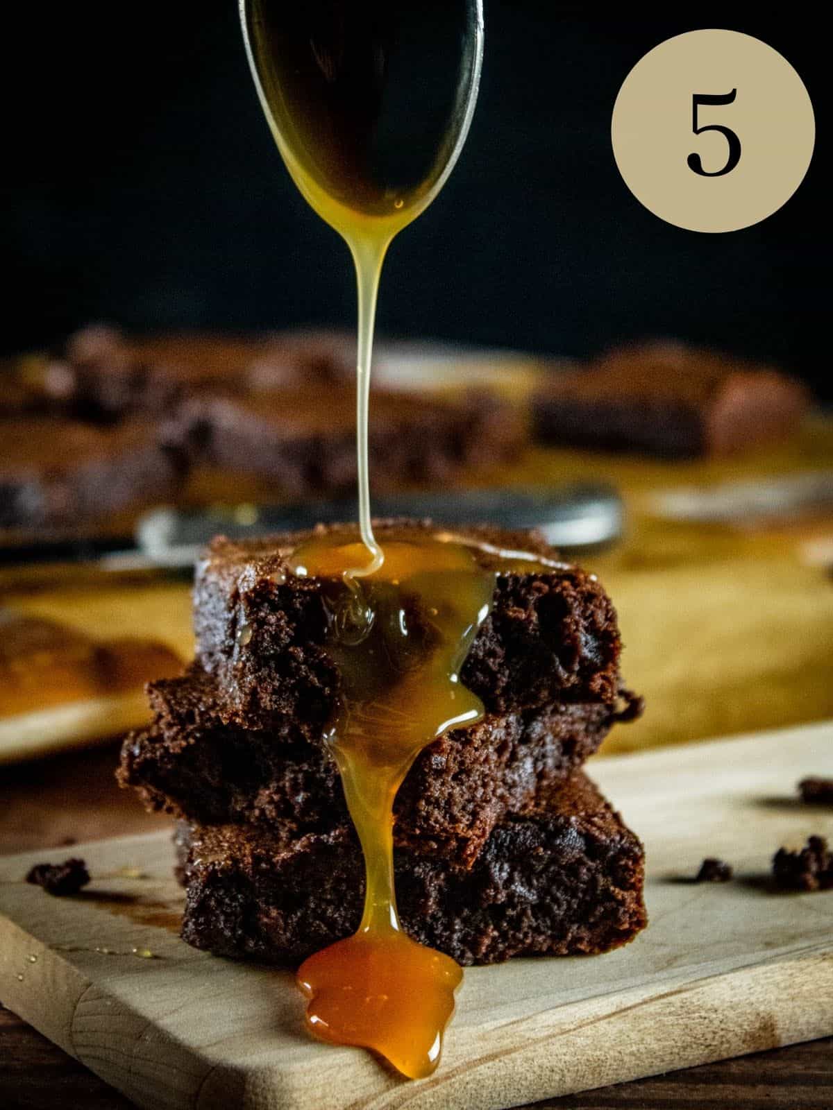 spoon drizzling caramel sauce over a stack of three brownies on a wooden tray