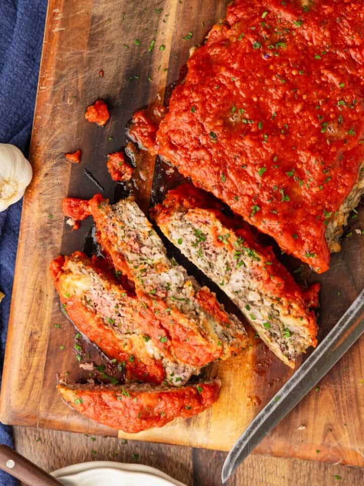 Meatloaf topped with marinara partially sliced on a cutting board with a knife.