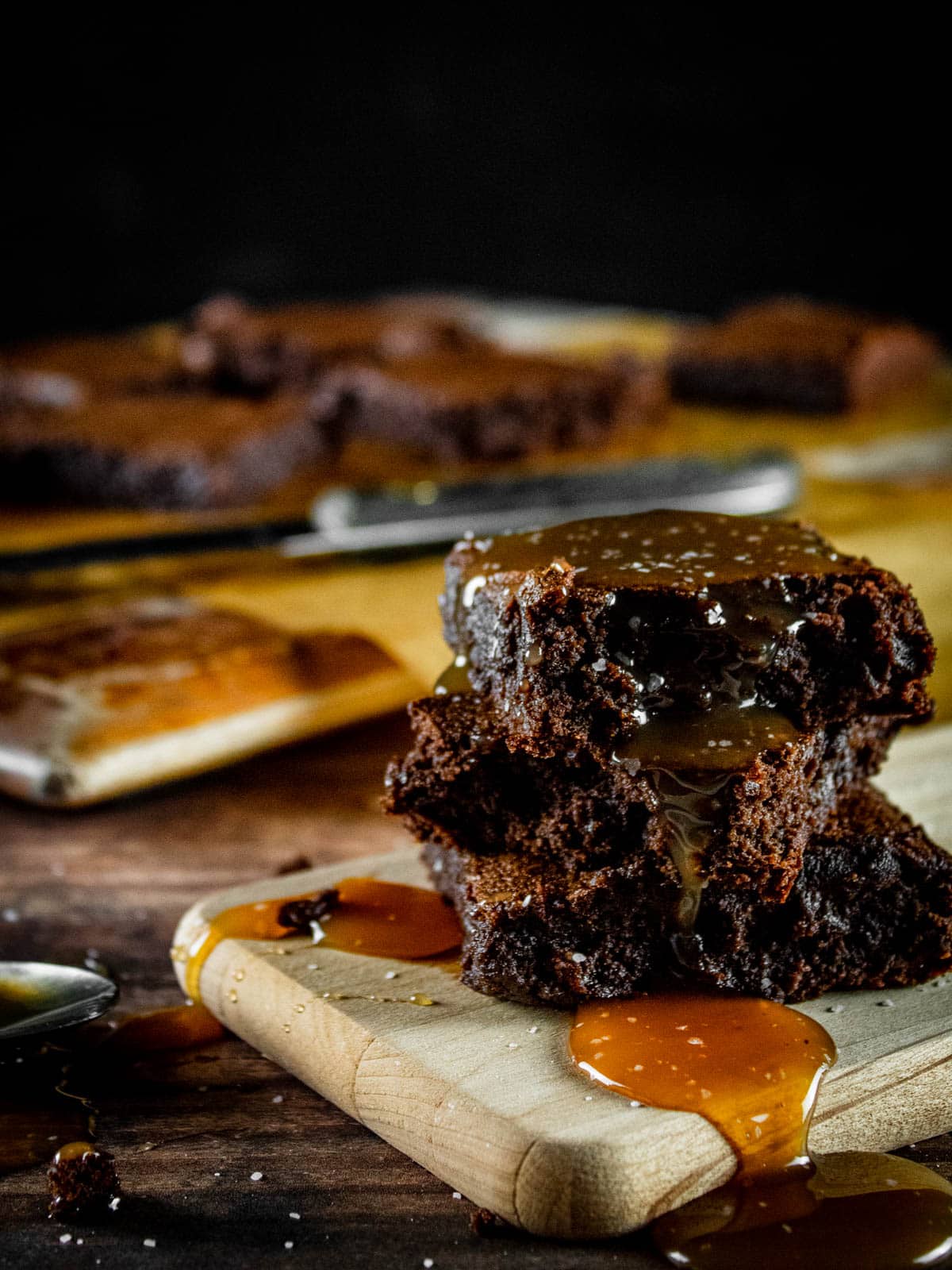stack of three brownies on a wooden tray with caramel sauce and sea salt on them