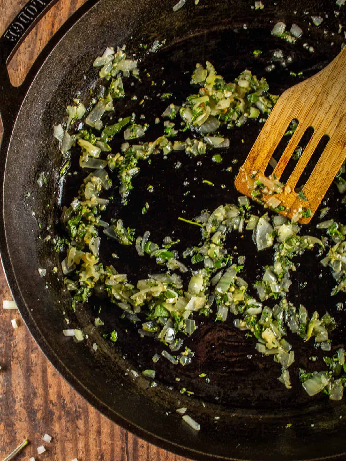 wooden spatula stirring herbs and onions in a cast iron skillet
