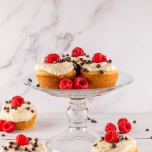 frosted cannoli cupcakes on a glass cake stand with fresh raspberries and chocolate chips.