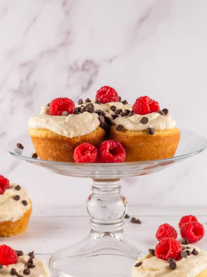 frosted cannoli cupcakes on a glass cake stand with fresh raspberries and chocolate chips.