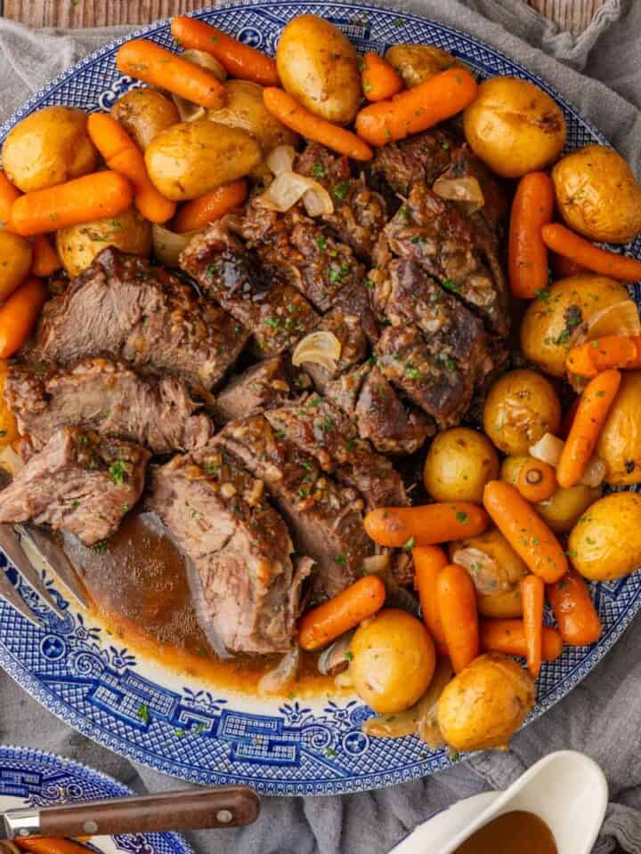 sliced pot roast on a round blue and white platter with baby yellow potatoes and baby carrots with gravy in a dish next to it.