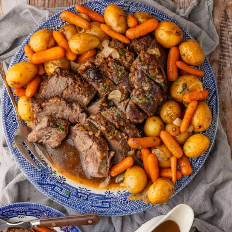 sliced pot roast on a round blue and white platter with baby yellow potatoes and baby carrots with gravy in a dish next to it.