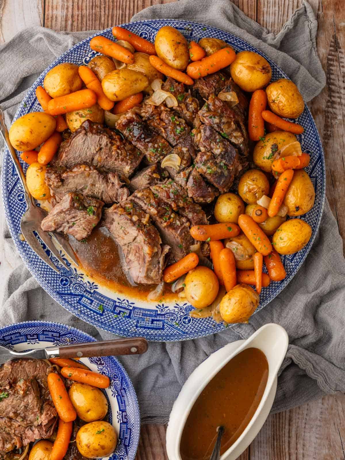 sliced pot roast with potatoes and carrots on a platter and a plate with a gravy boat next to it.