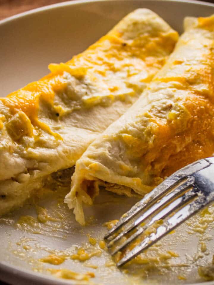 two cheese covered chicken enchiladas with white sauce on a plate with a fork with some bites taken from it.