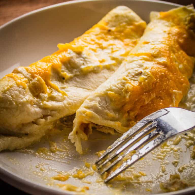 two cheese covered chicken enchiladas with white sauce on a plate with a fork with some bites taken from it.