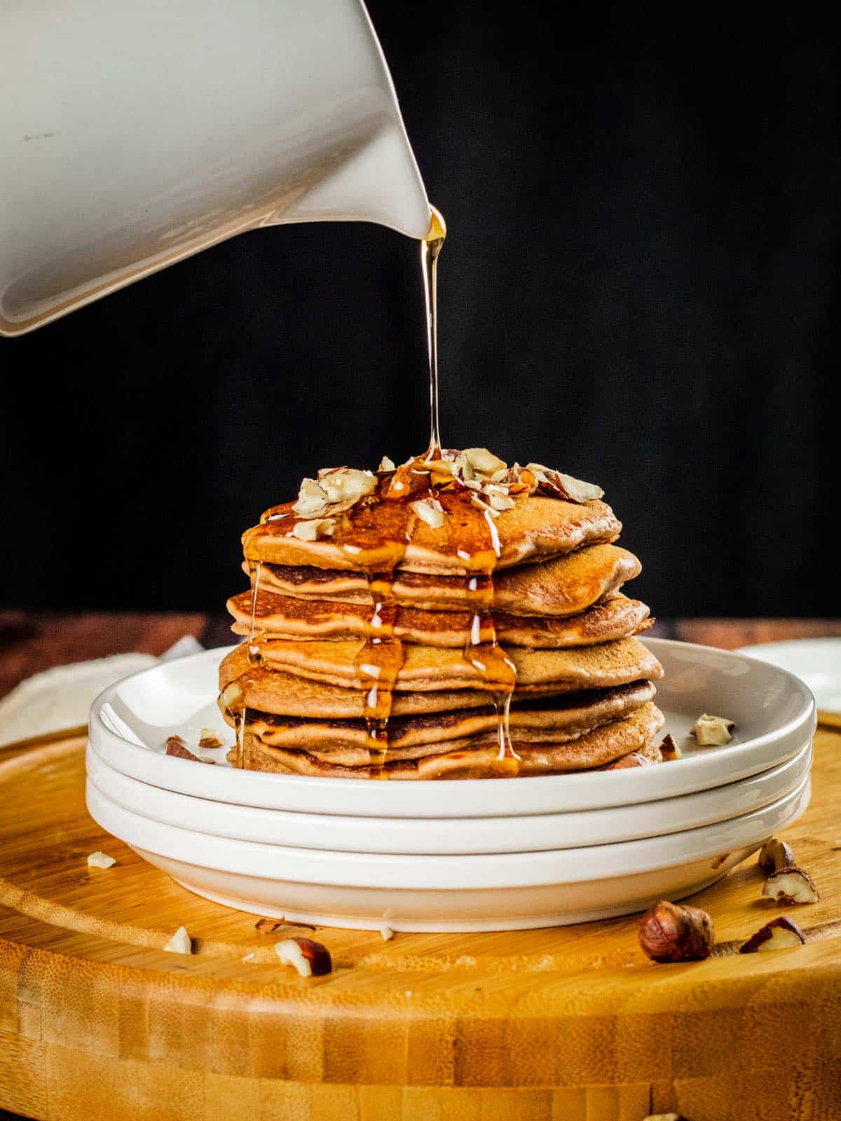 white pitcher pouring syrup over a stack of nutella pancakes on a stack of white plates with chopped hazelnuts.