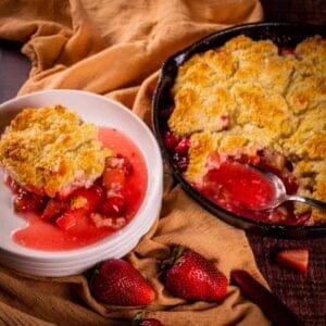 skillet with strawberry rhubarb cobbler and a serving on a white plate