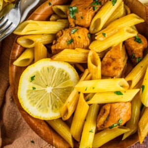 penne pasta in a wooden bowl with chicken, fresh parsley a fork and lemon slices