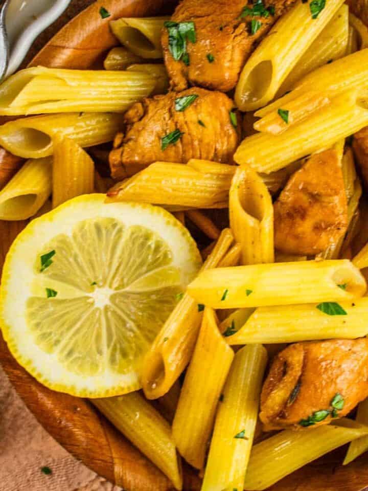 penne pasta in a wooden bowl with chicken, fresh parsley a fork and lemon slices
