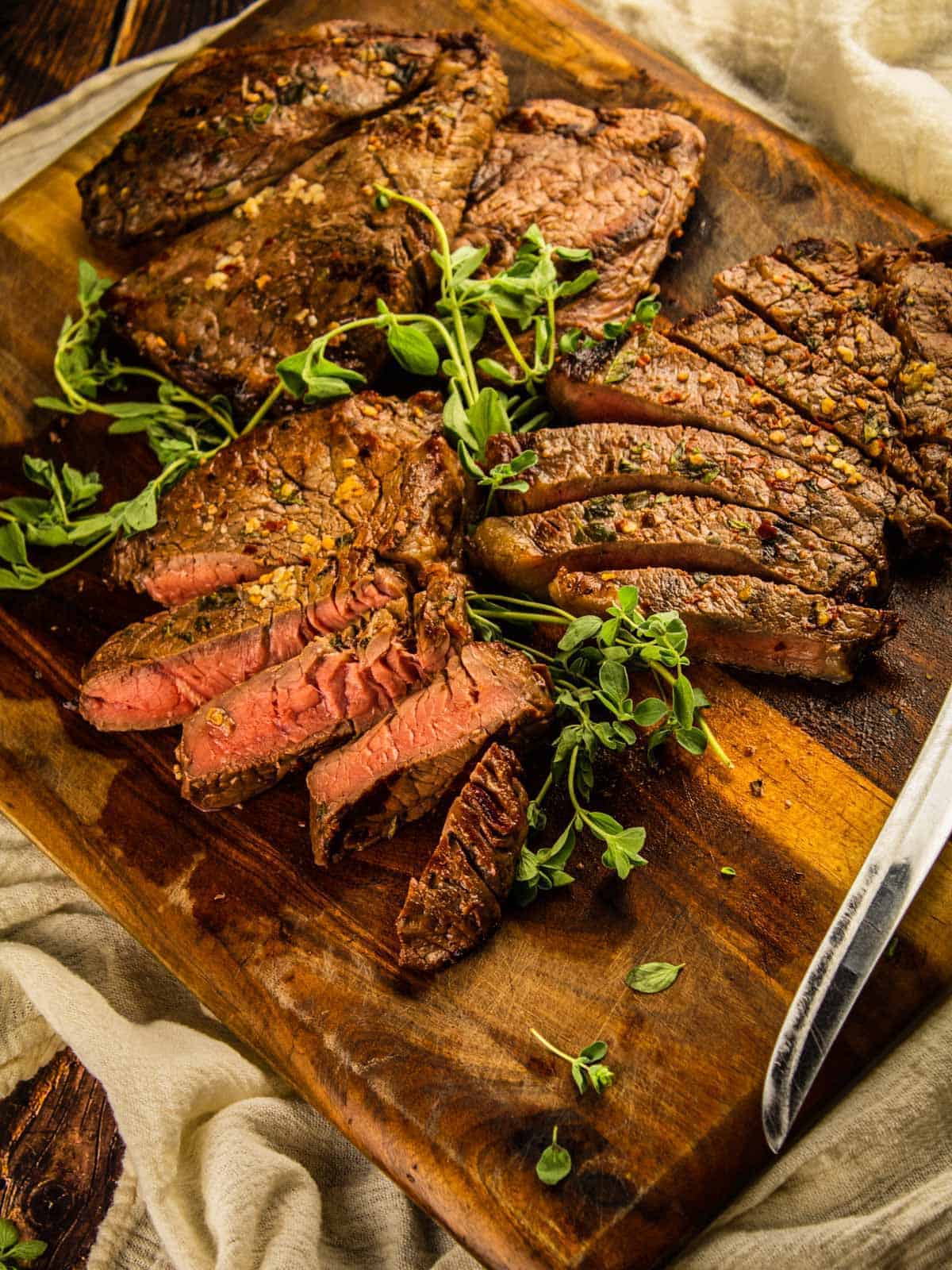 grilled steak sliced on a cutting board with fresh oregano and a knife.
