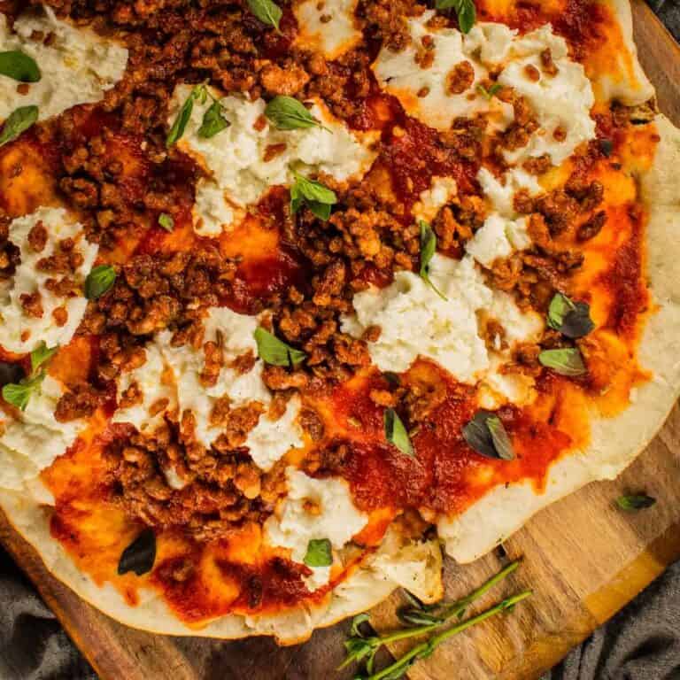 grilled pizza on a wooden cutting board topped with marinara, ricotta, sausage and fresh oregano leaves