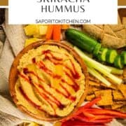 hummus in a wooden bowl drizzled with sriracha and next to crackers and vegetables