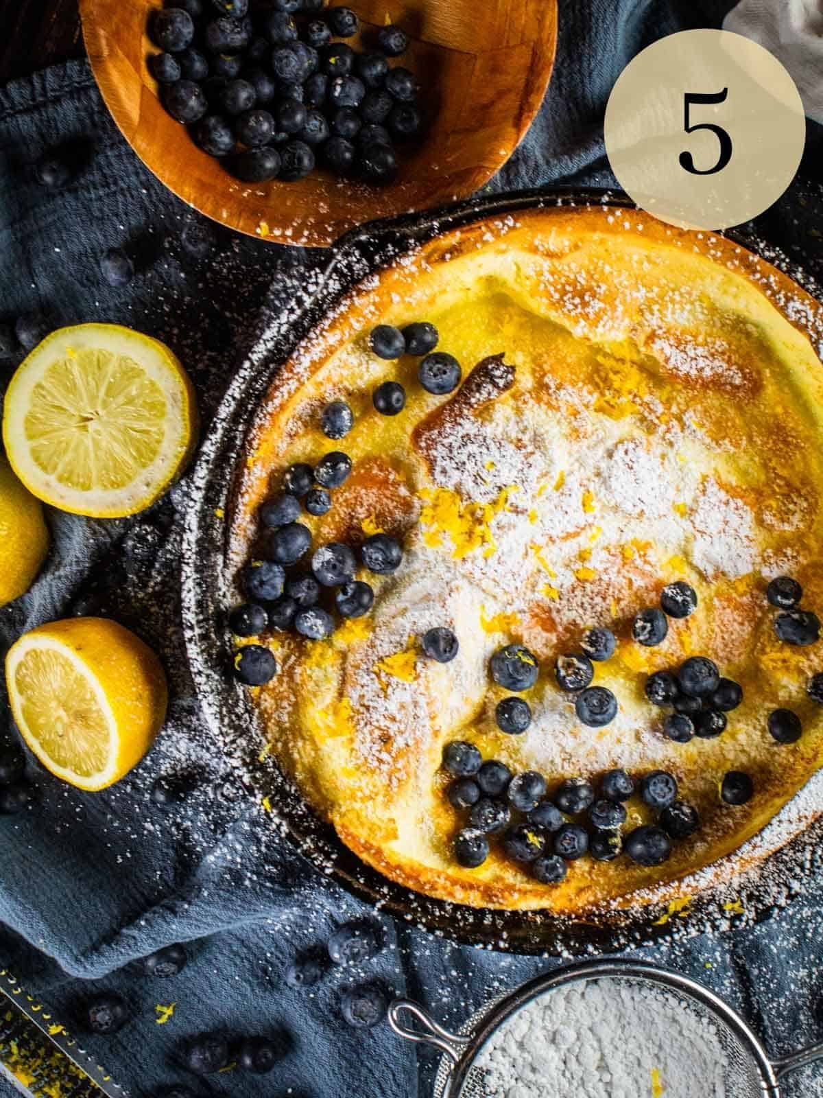 dutch baby german pancake in a skillet topped with powdered sugar, blueberries and lemon zest