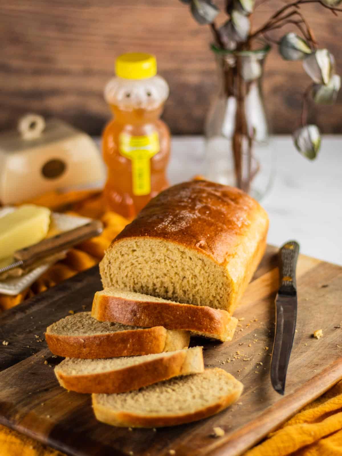 loaf of bread sliced on a wooden cutting board with knife, butter and honey