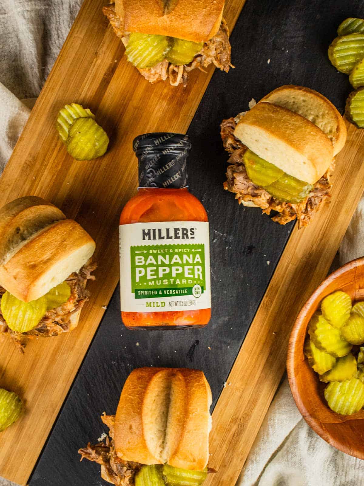 pulled pork on slider buns with pickles around a bottle of millers mustard on a cutting board