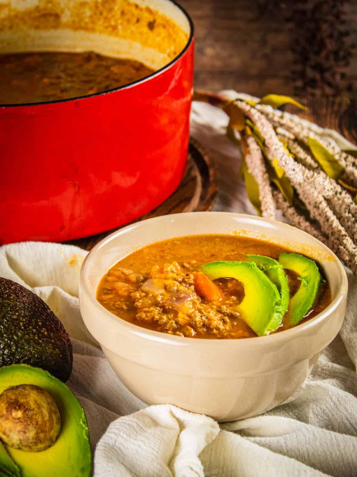 bowl of sweet potato turkey chili topped with sliced avocado with avocados next to it and a red pot in the background