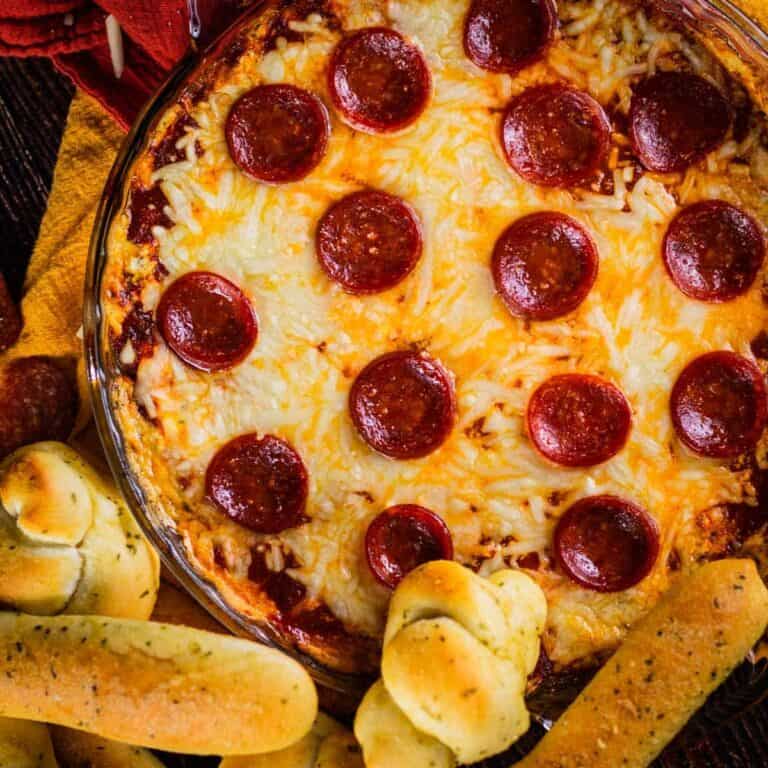 baked cheesy pizza dip topped with pepperoni slices next to soft breadsticks