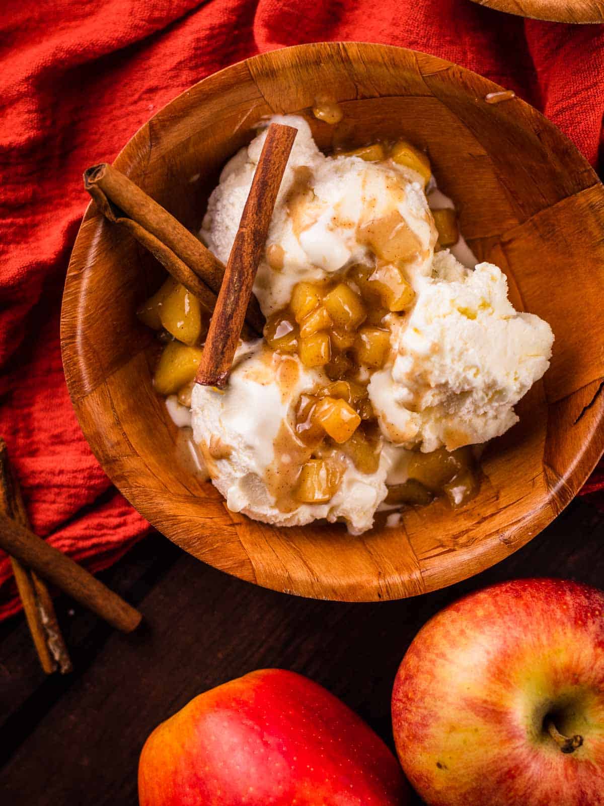 wooden bowl filled with ice cream, caramel apple topping and two cinnamon sticks next to apples