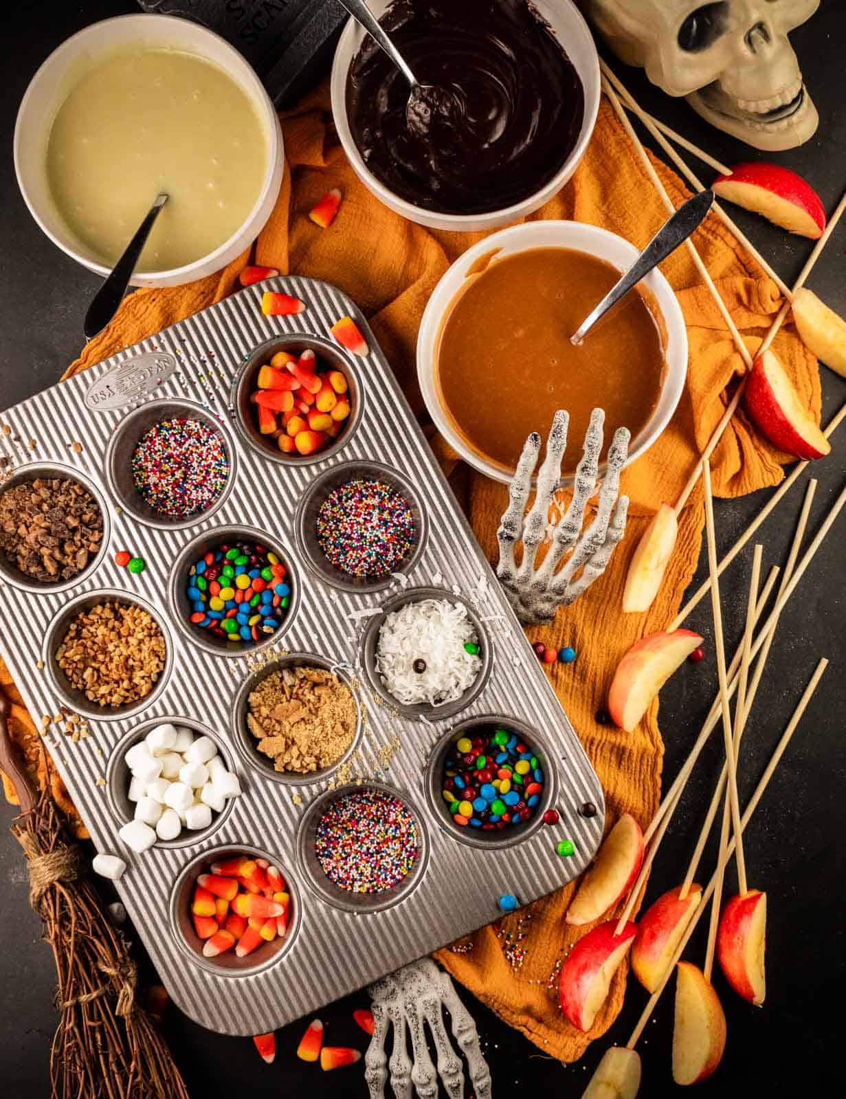 muffin tin with assorted candies, white and milk chocolate and caramel dip, apple slices on skewers