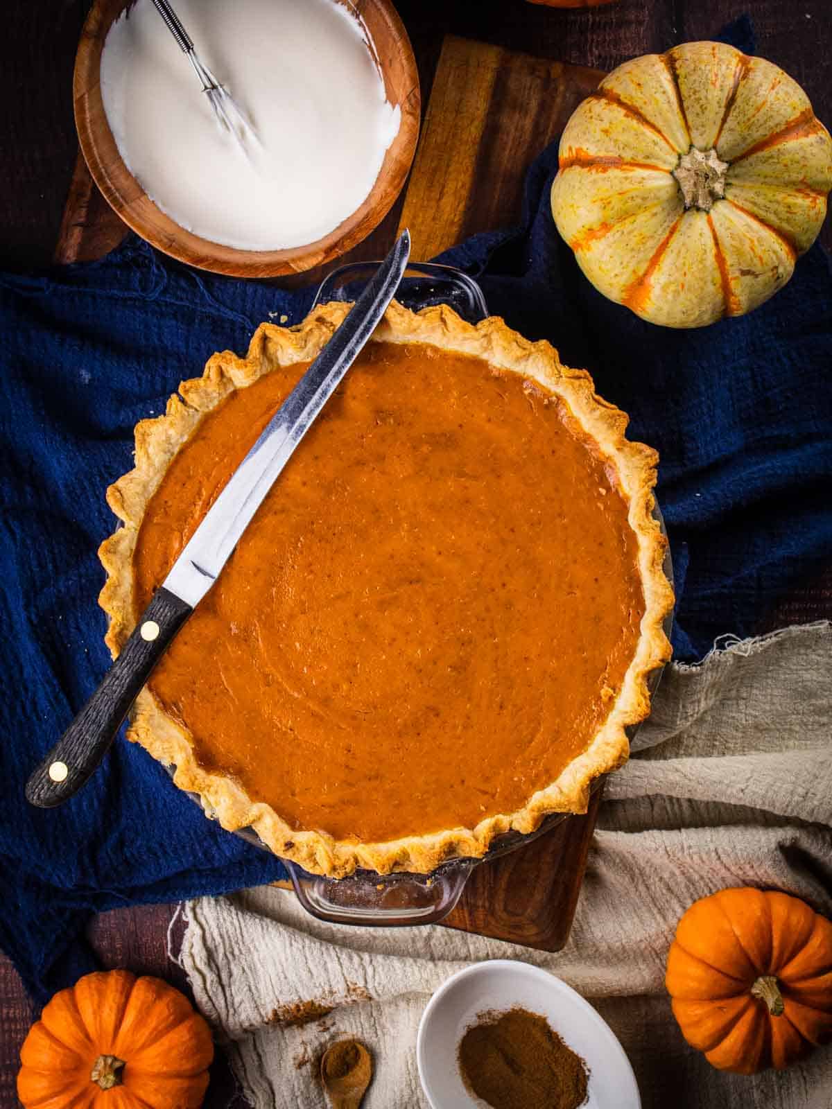 pumpkin pie with a knife on top, a bowl of whipped cream and fresh pumpkins around