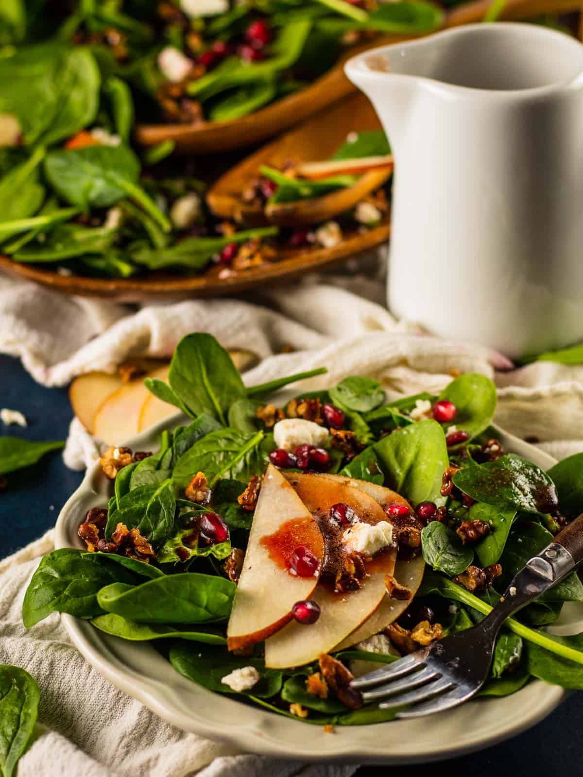 spinach on a plate with pears, pomegranate, candied pecans and gorgonzola and dressing pitcher behind it