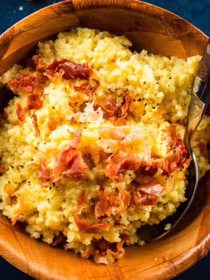 wooden bowl filled with cheesy pastina topped with crispy prosciutto and a spoon sticking in it