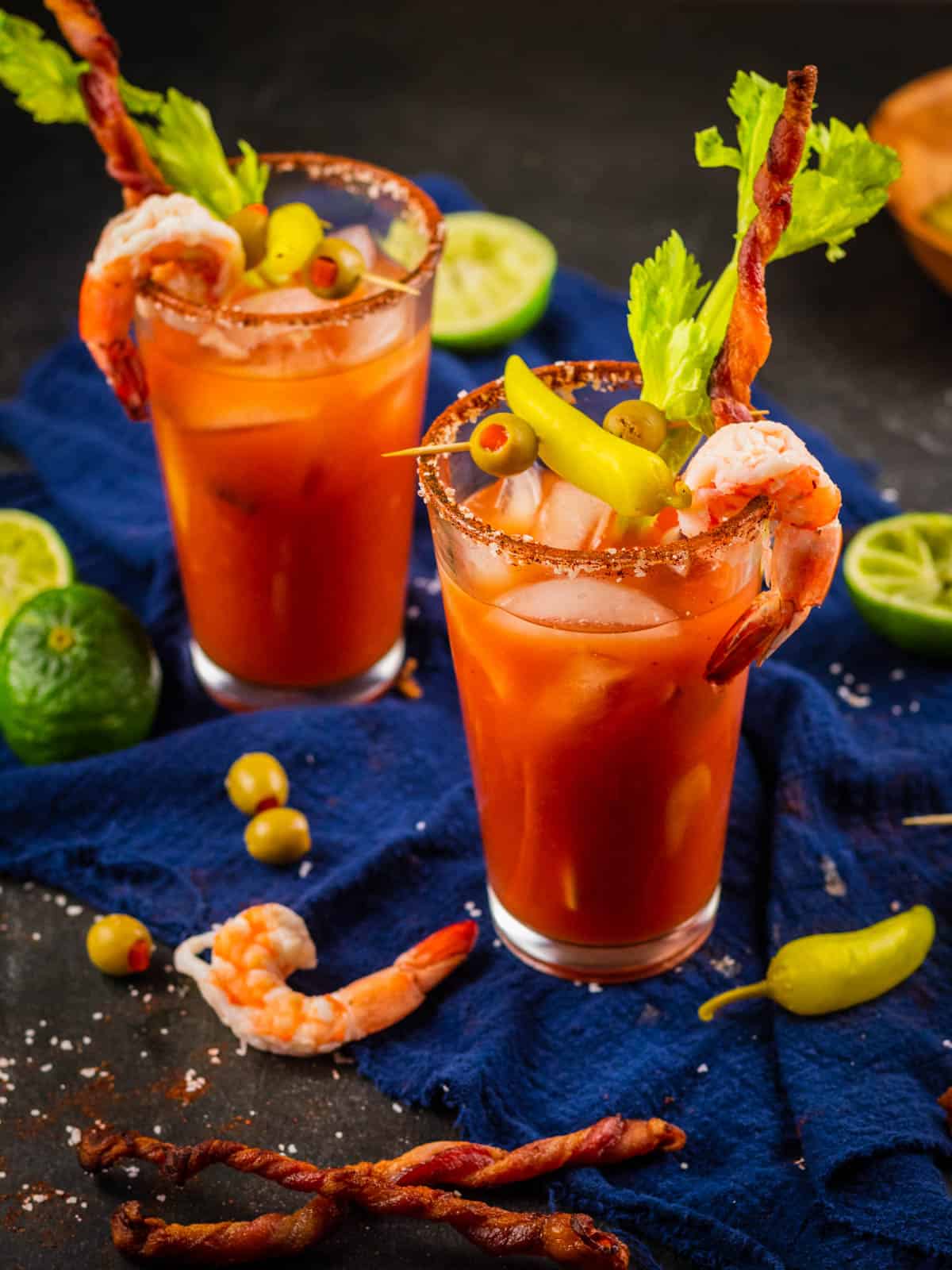 two bloody mary cocktails garnished with peppers, celery, shrimp, olives and bacon.