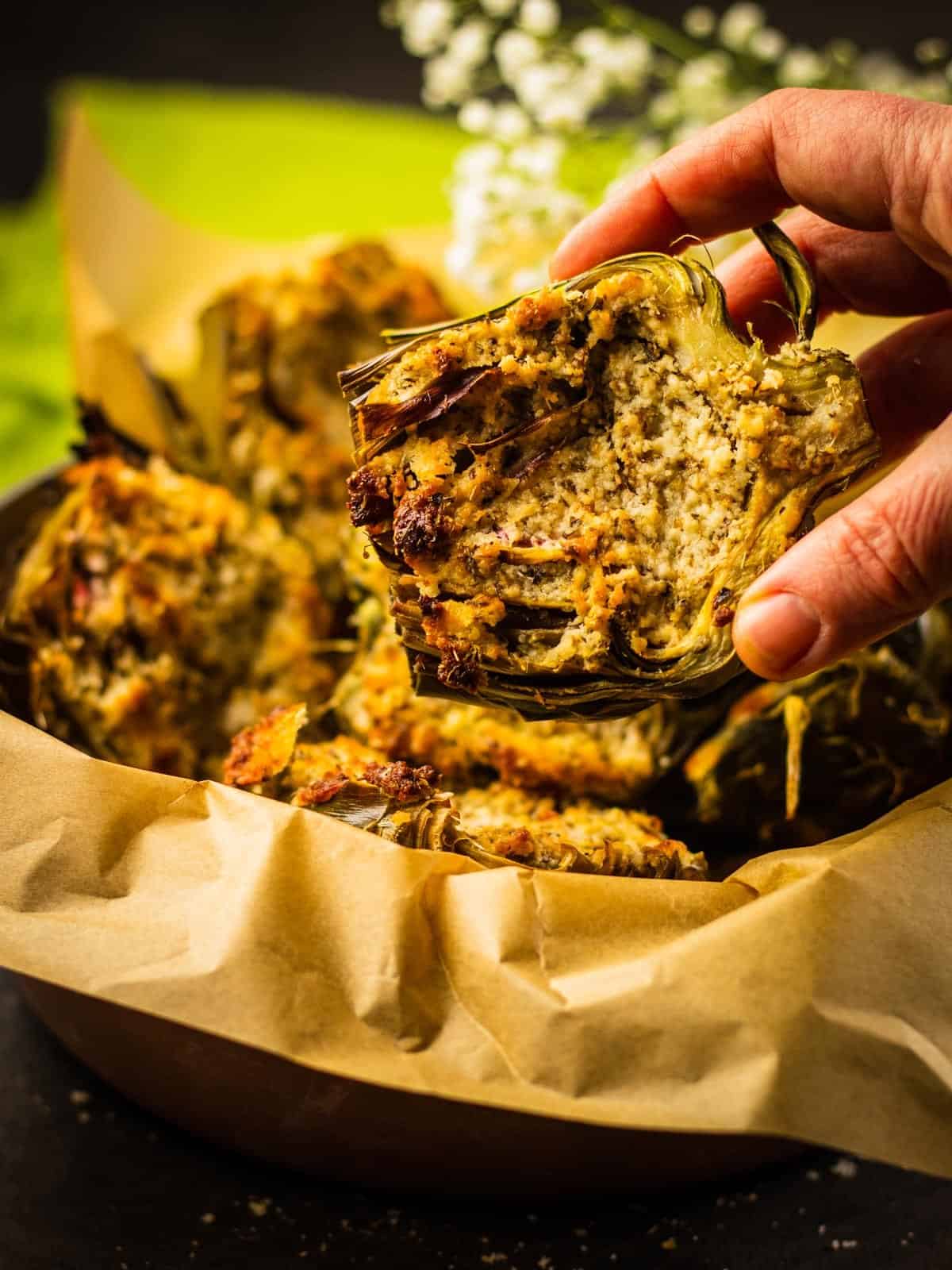 hand grabbing a parmesan crusted roasted artichoke half out of a basket lined with parchment