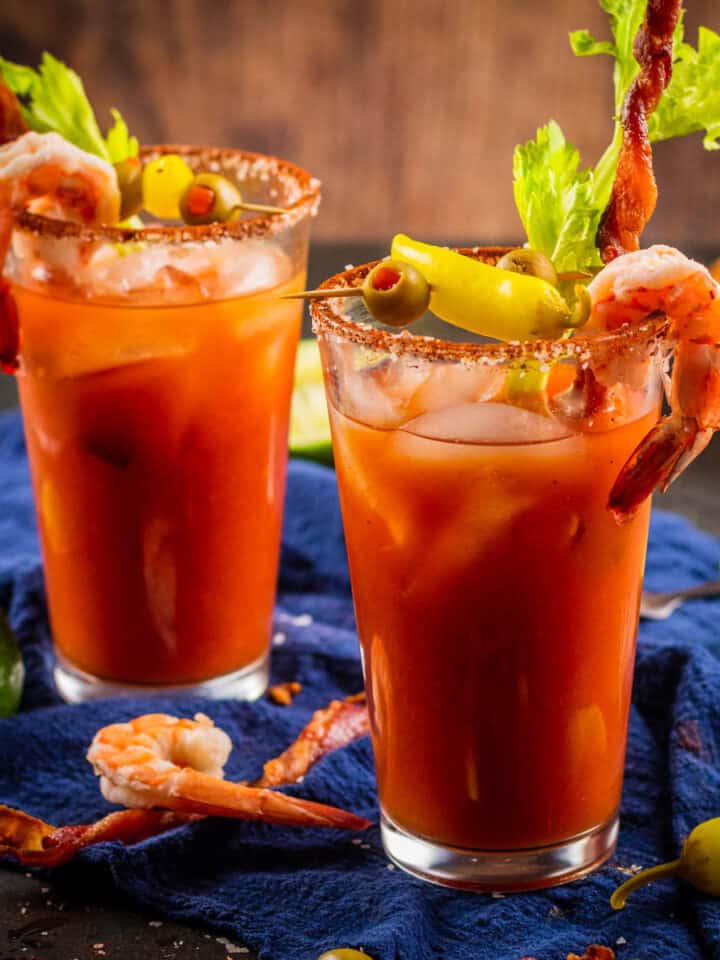two bloody marys garnished with shrimp, bacon, celery, olives and peppers