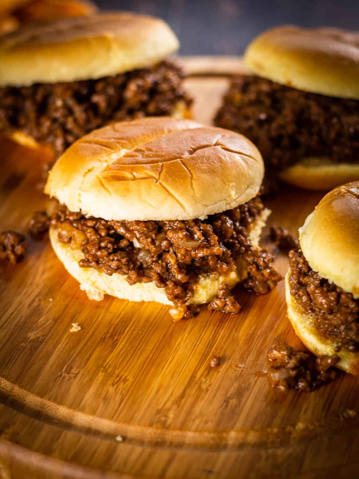 sloppy joe sandwiches on hamburger buns on a round wooden tray with meat spilling out.
