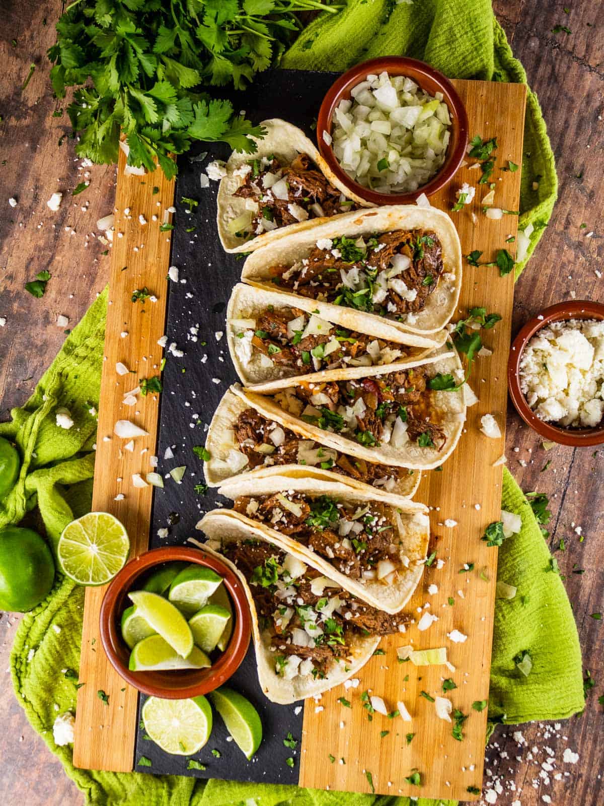 barbacoa beef tacos lined up on wooden tray with lime slices, queso fresco, onions and cilantro