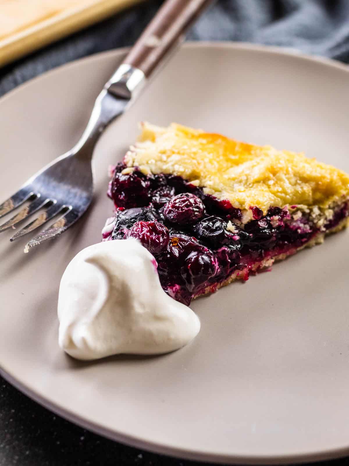 slice of blueberry crostata with a dollop of whipped cream on a gray plate with a fork.