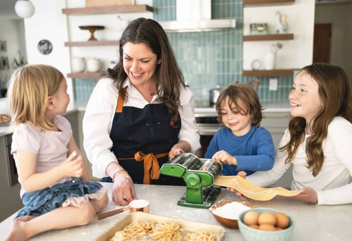 a woman and three children in a kitchen rolling and cutting pasta.