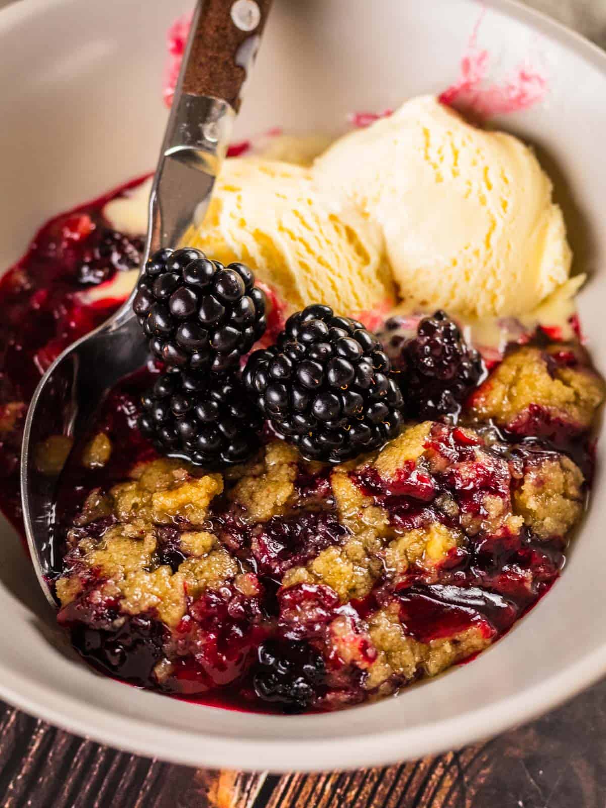 bowl of blackberry crumble topped with ice cream and fresh blackberries with a spoon in it.