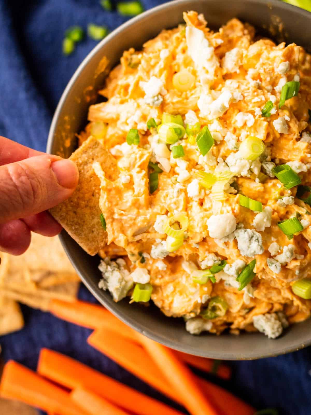 hand dipping cracker into buffalo chicken dip topped with blue cheese and green onions.