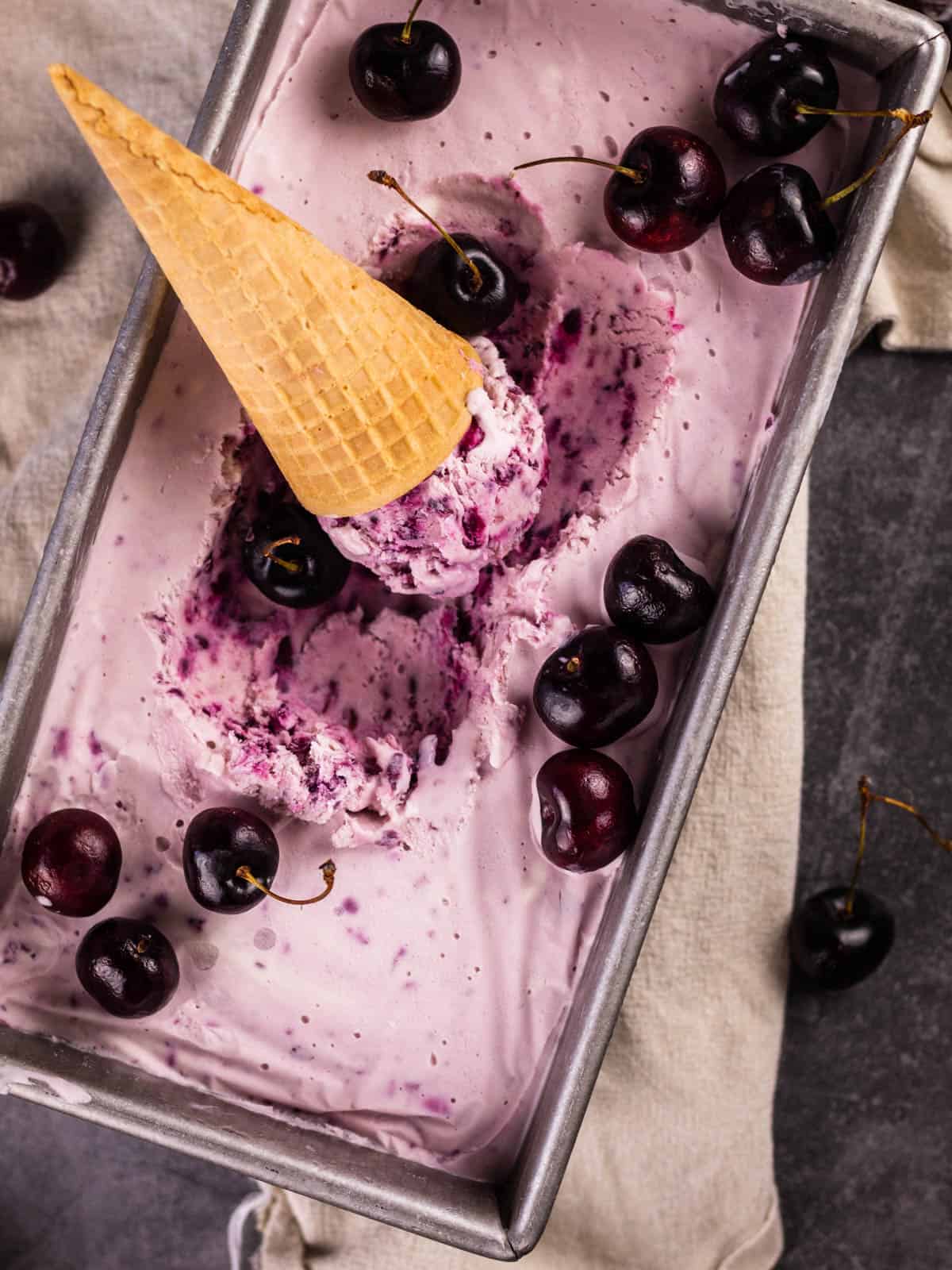 pan of black cherry ice cream with cherries and a ice cream cone sitting in it.