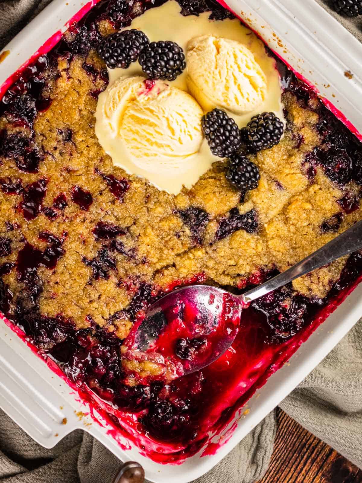 blackberry crumble in white square ceramic dish topped with ice cream with spoon scooping it out.