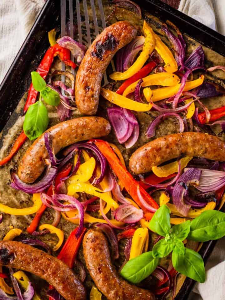 baked sausage, peppers and onions on a sheet pan with fresh basil sprigs around.