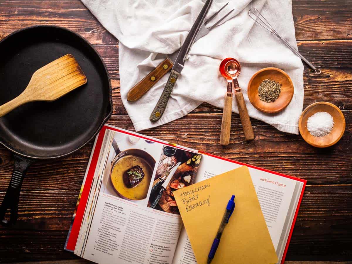cookbook with a shopping list and pen next to cooking tools and a skillet.