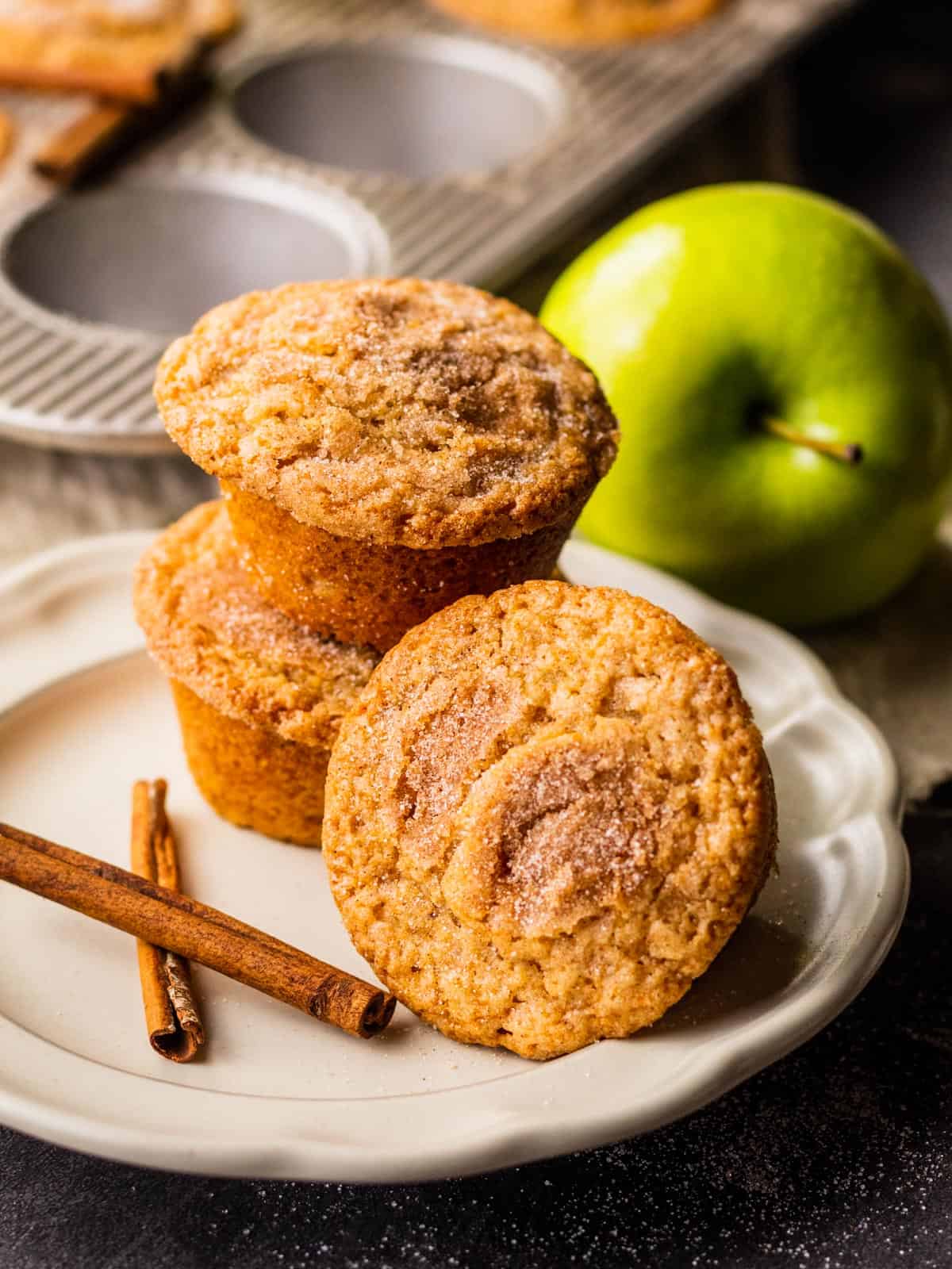 cinnamon sugar dusted muffins stacked on a plate next to two cinnamon sticks and green apple.