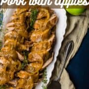 sliced pork tenderloin with gravy on a platter with apples and fresh herbs around.