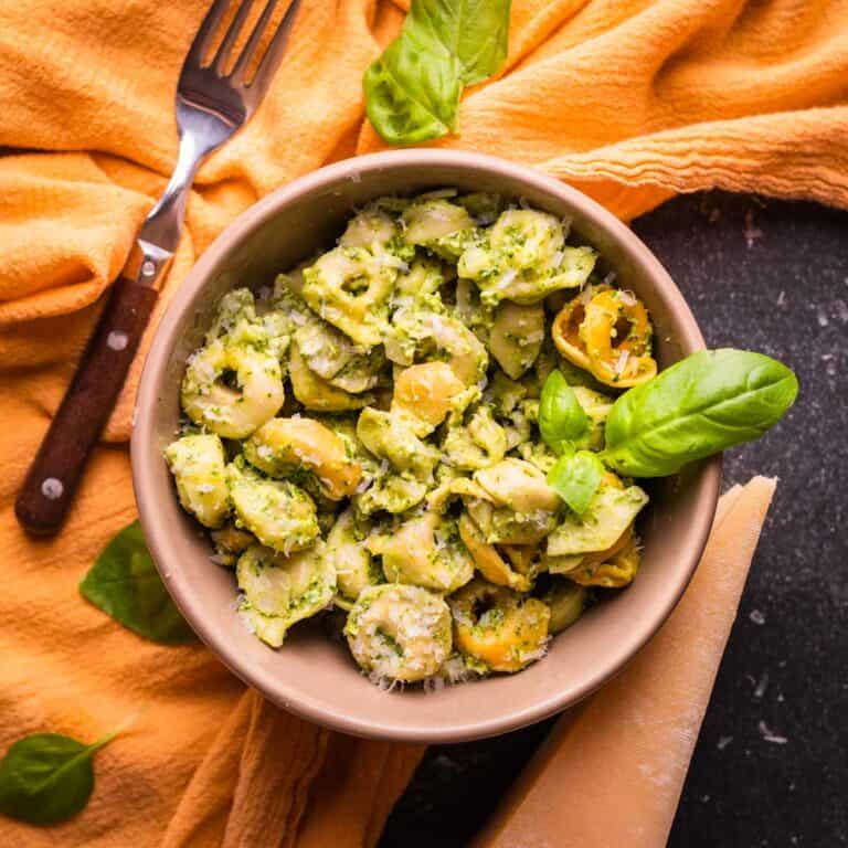 bowl of colored tortellini with creamy pesto sauce and a fresh basil leaf garnish.