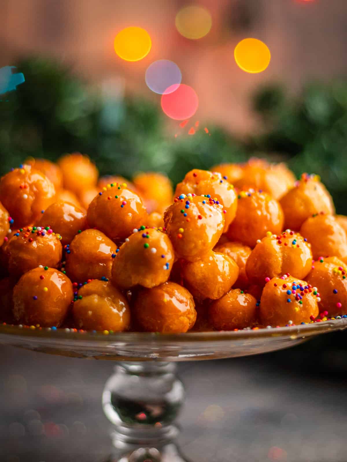 italian honey balls with rainbow sprinkles on a glass cake stand with christmas lights behind.